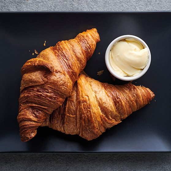 Dwa złociste, świeżo upieczone croissanty na czarnym talerzu z miseczką masła – przygotowane dzięki CHEF'S COMBI od HOBART, zapewniające idealną teksturę i zarumienienie.