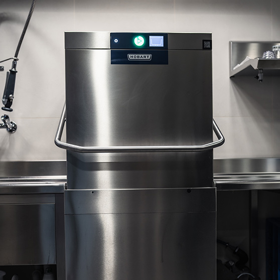 Widok z przodu na zmywarkę kapturową HOBART AUP w profesjonalnej kuchni, z podświetlonym panelem sterowania i stalową obudową, zapewniającą szybkie i niezawodne mycie w godzinach szczytu oraz zintegrowaną z systemem Creatus w celu optymalizacji zużycia energii.