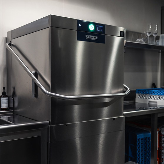 Widok z boku na zmywarkę kapturową HOBART AUP w profesjonalnej strefie zmywania, z otaczającymi ją stalowymi blatami i półkami, ukazujący jej solidną konstrukcję, cichą pracę oraz wydajność w szczytowym obciążeniu dzięki integracji z systemem Creatus.