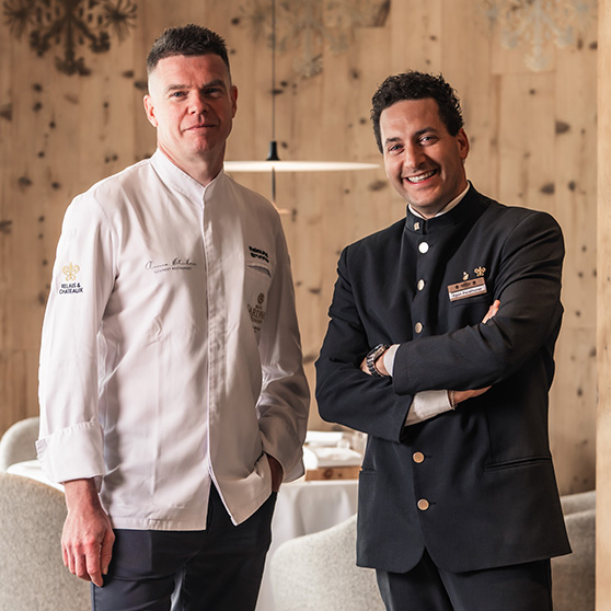 Dwie osoby ubrane w formalny strój związany z branżą hotelarską stoją w restauracji z drewnianymi ścianami i przygotowanymi stołami, co nawiązuje do tekstu opisującego znaczenie bezpiecznych procesów i niezawodnej technologii zmywania, takiej jak systemy HOBART.