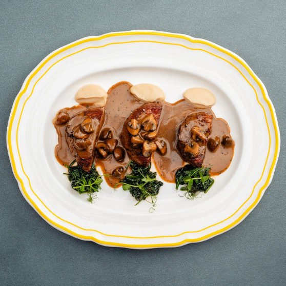 Biały talerz z żółtym brzegiem z trzema kawałkami mięsa w sosie śmietanowo-grzybowym, udekorowany pieczarkami, jasnym puree i zielonymi ziołami, elegancko podany na szarym stole.