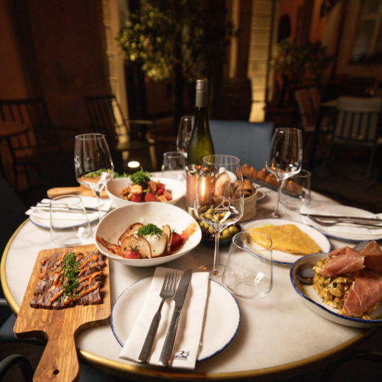 Stół restauracyjny na zewnątrz starannie nakryty kilkoma daniami, przystawkami, pieczywem, butelką wina i różnymi kieliszkami.
