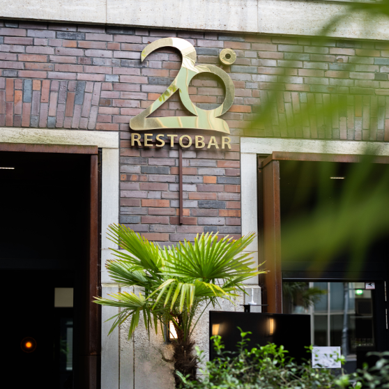 Widok zewnętrzny budynku z cegły z złotym szyldem 20° RESTOBAR nad wejściem, otoczony zielonymi roślinami.