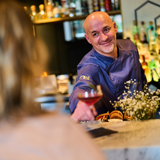 Barman podaje napój gościowi przez blat baru, a w tle widać butelki i półki barowe.