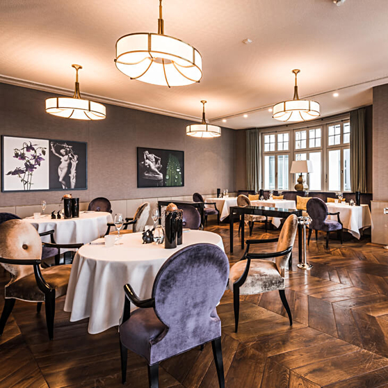Eleganckie wnętrze restauracji fine‑dining z okrągłymi stołami nakrytymi na biało, tapicerowanymi krzesłami i ciepłym oświetleniem tworzącym ekskluzywny klimat.