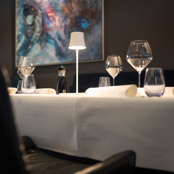 Zbliżenie na elegancko nakryty stół w restauracji RIVA z wyrafinowanymi kieliszkami, białymi serwetami i nowoczesnym oświetleniem.